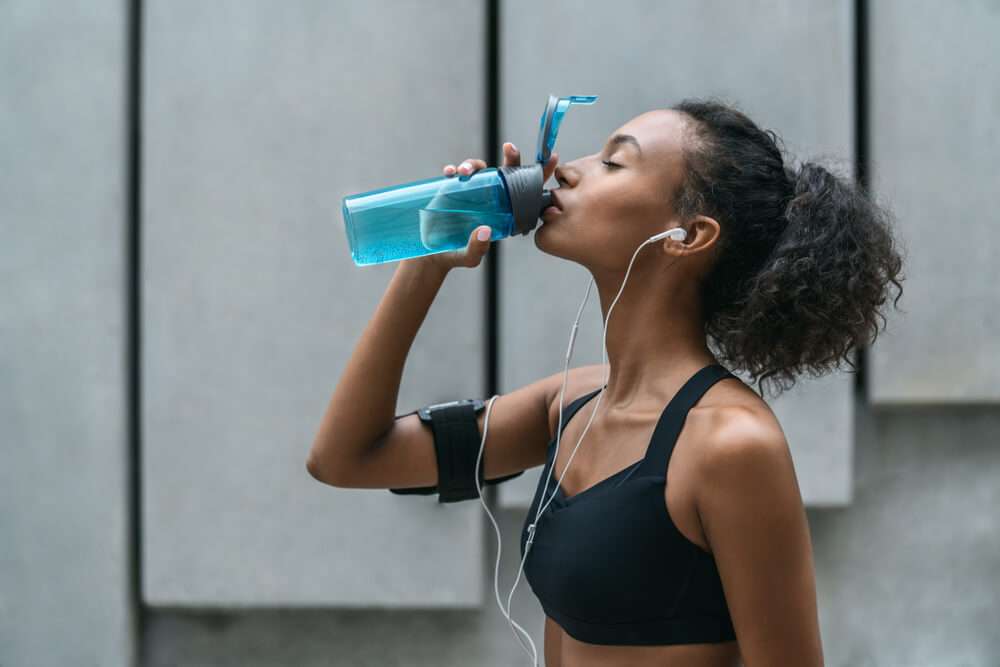 Fitness woman drinking water from a bottle after workout standing by a grey wall
