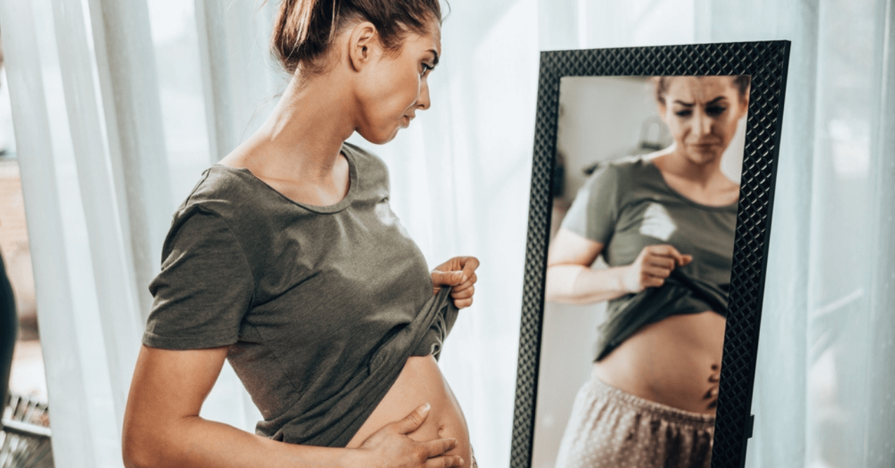 Unhappy woman standing in front of a mirror and holding her stomach while suffering from bloating