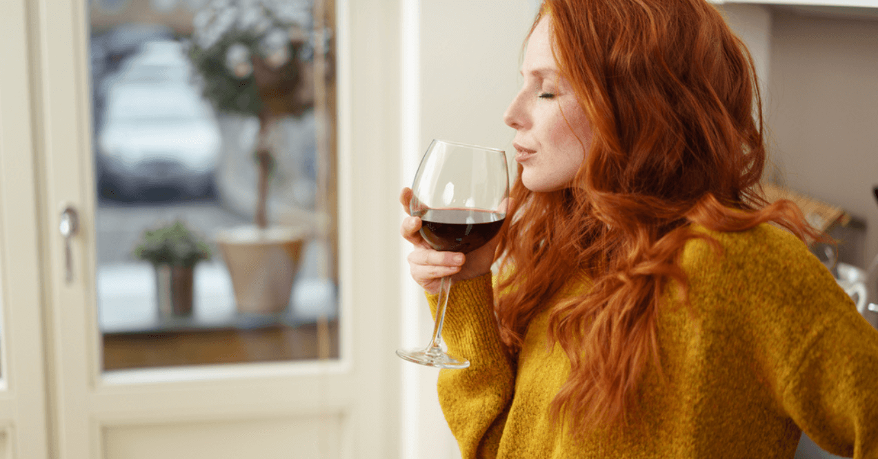 Young redhead woman standing in her apartment sipping a glass of red wine with her eyes closed in pleasure