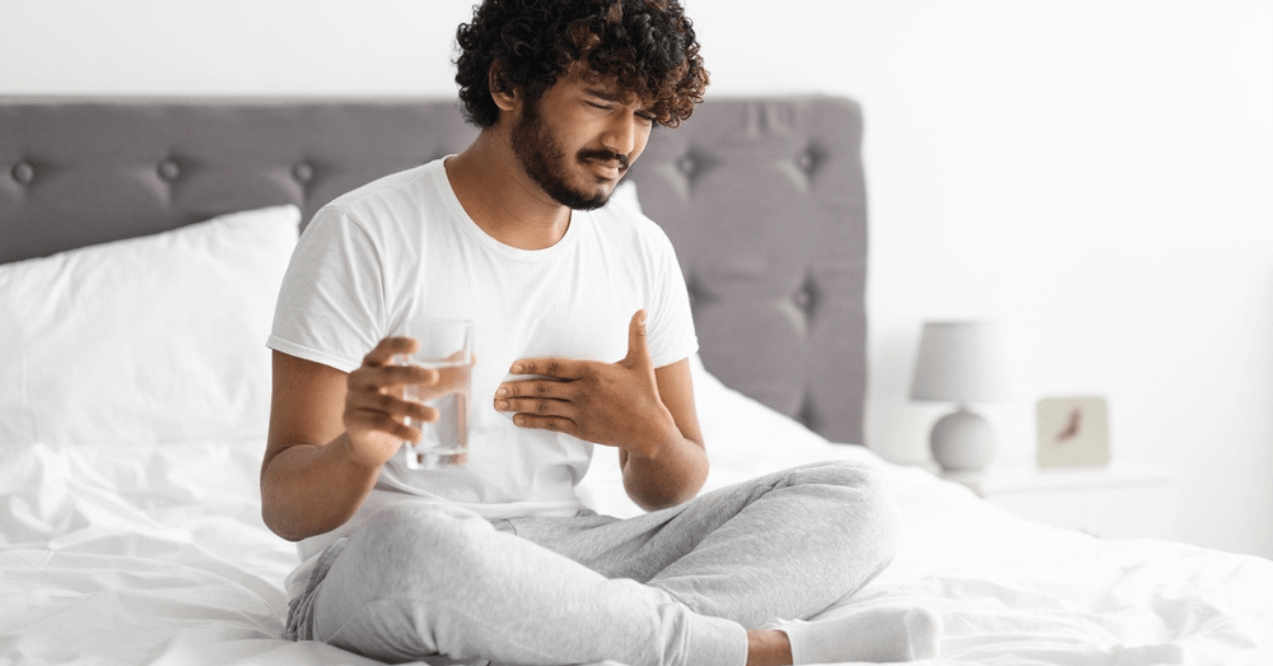 Unhappy millennial indian guy suffering from heartburn, waking up in morning, sitting on bed and holding glass of water