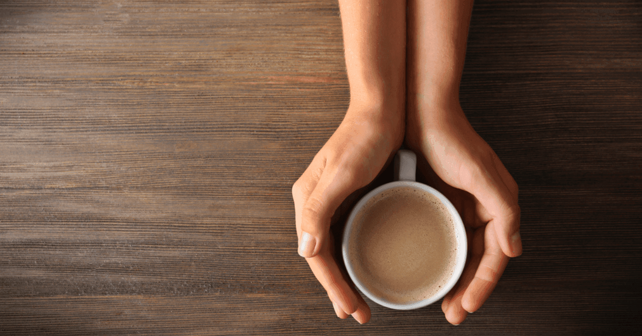 Female hands holding a cup of coffee with foam over wooden table, top view