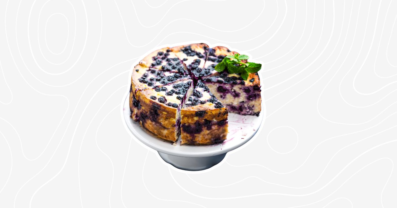 A round blueberry coffee cake topped with fresh blueberries and a mint garnish, served on a cake stand.