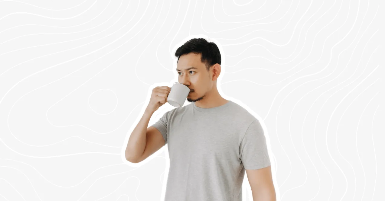 A man in a gray t-shirt drinking coffee from a white mug, with a neutral expression on his face, against a minimalistic white background.